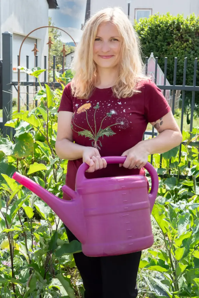 Frau in rotem T-Shirt, stehend im Garten beim gießen der Pflanzen mit einer pink-farbigen Gießkanne.