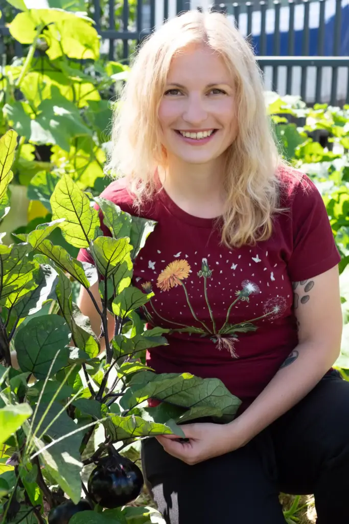 Blonde Frau kniend im Garten mit einem roten T-Shirt und präsentiert ihre angebauten Pflanzen.