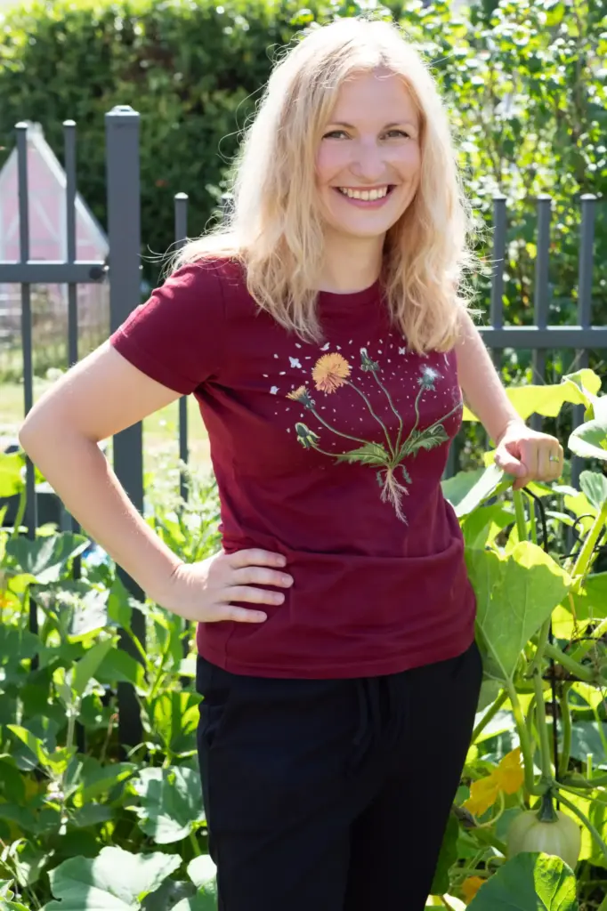 Blonde Frau steht im Garten, trägt rotes T-Shirt und stämmt ihre Hände in die Taille. Bild verdeutlicht eine klare auffordernde Handlung, um Kontakt aufzunehmen bei Fragen oder bei Interesse zum Garten-Coaching, zu den Online-Kurse oder zu allgemeinen Fragen zur Gartenberatung oder Balkonberatung.