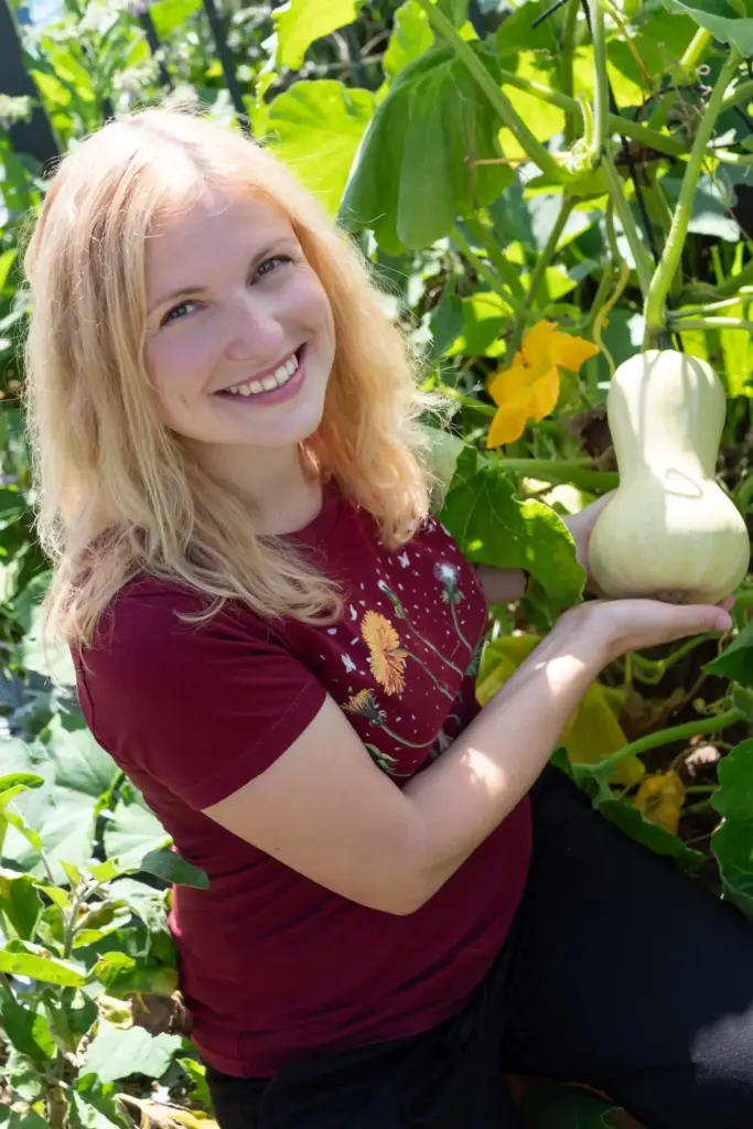 Blonde Frau sitzt kniend auf dem Boden und präsentiert ein Gemüse in ihrem Garten.