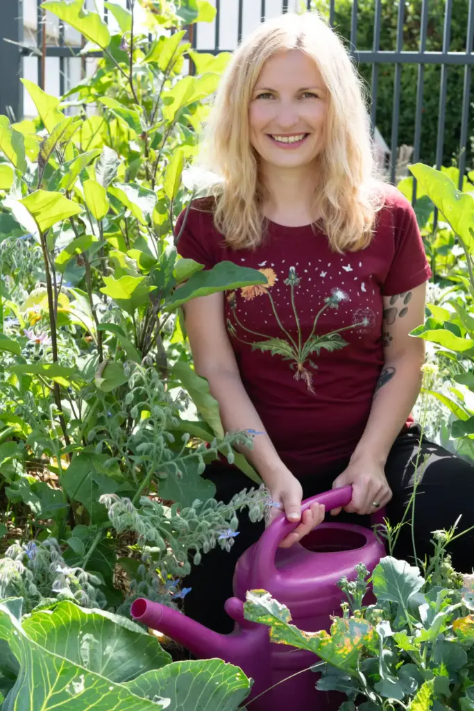 Frau kniet im Garten mit einer pinken Gießkanne und versorgt die Kräuter und Pflanzen. Sie schaut in die Kamera und lächelt.