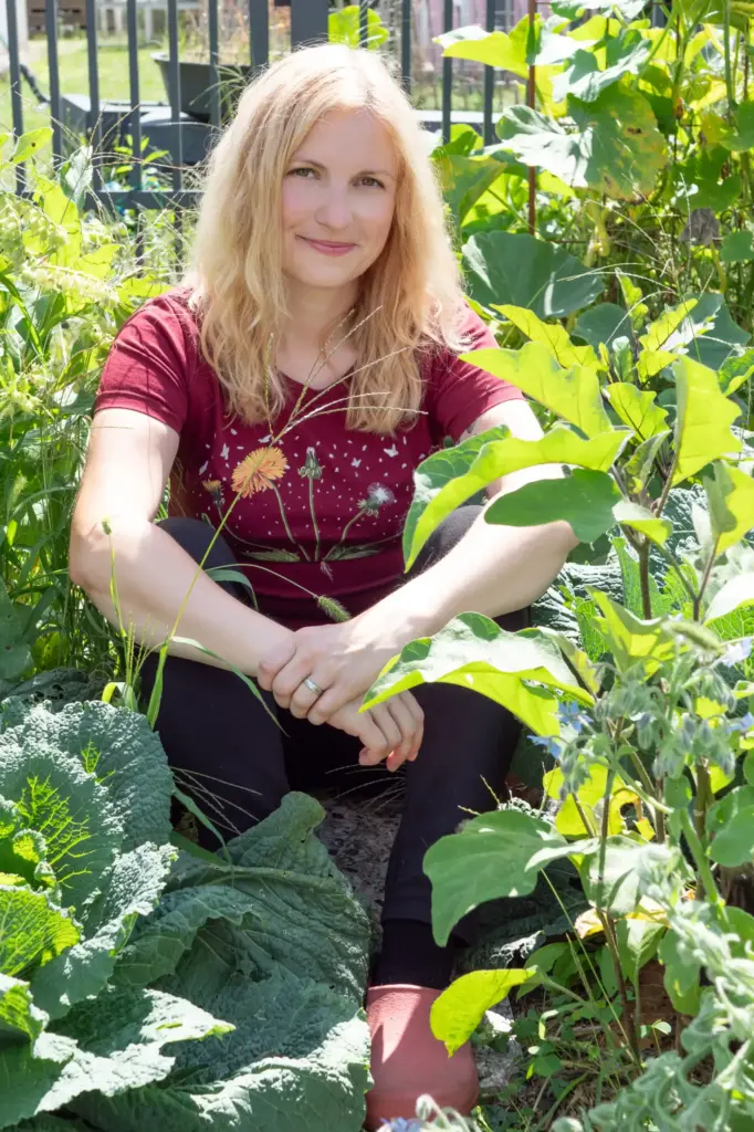 Blonde Frau sitzt entspannt im Garten mit überschlagenen Füßen und stützt sich auf ihren Knien ab. Sie schaut zurückhalten in die Kamera und lächelt.