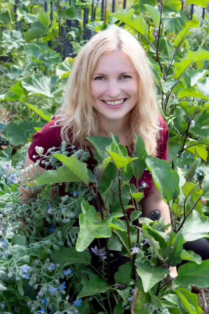 Julia sitzt kniend hinter Pflanzen und schaut in die Kamera.