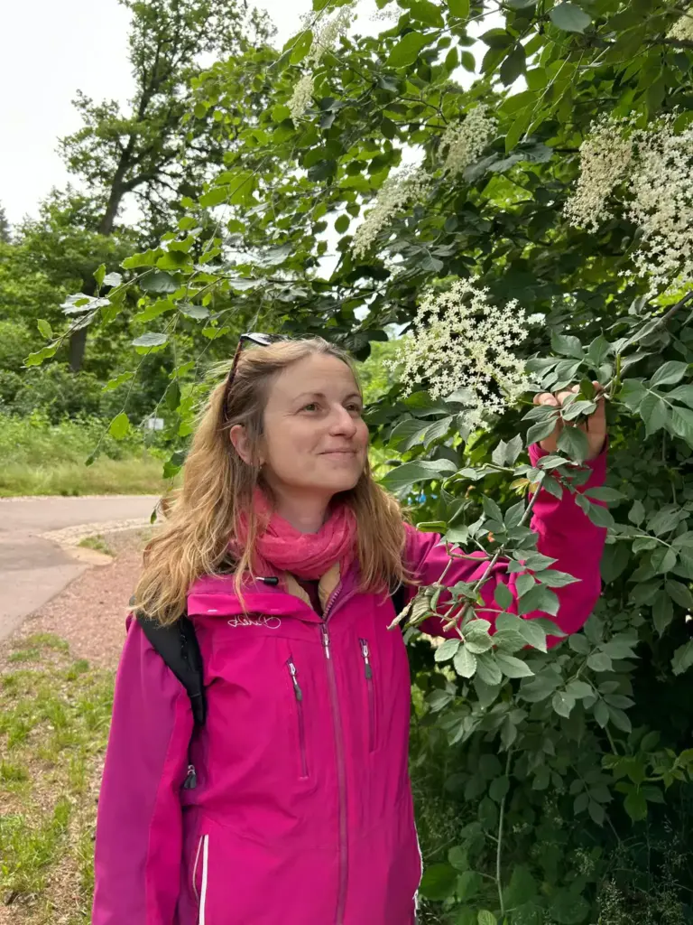 Julia steht neben einem Baum bei der Veranstaltung Kräuterwanderung und schaut die Blüten an. Neben den Kräuterwanderungen bietet Julia Junggesellenabschied, Kindergeburtstag und Workshops für Selbstversorger an, bei denen man gemeinsam auf Spaziergänge geht und die Wildkräuter erkundet.