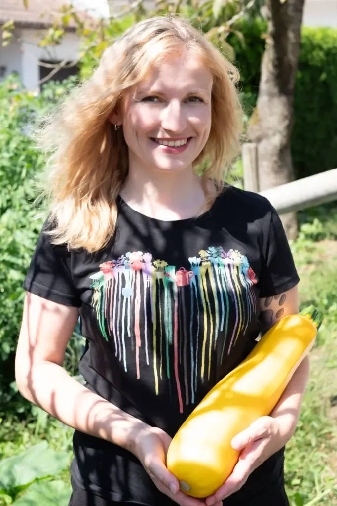 Frau mit schwarzem Tshirt hält große gelbe Zucchini in der Hand. Die Sonne scheint ihr in das Gesicht und die Haare werden nach hinten geweht.
