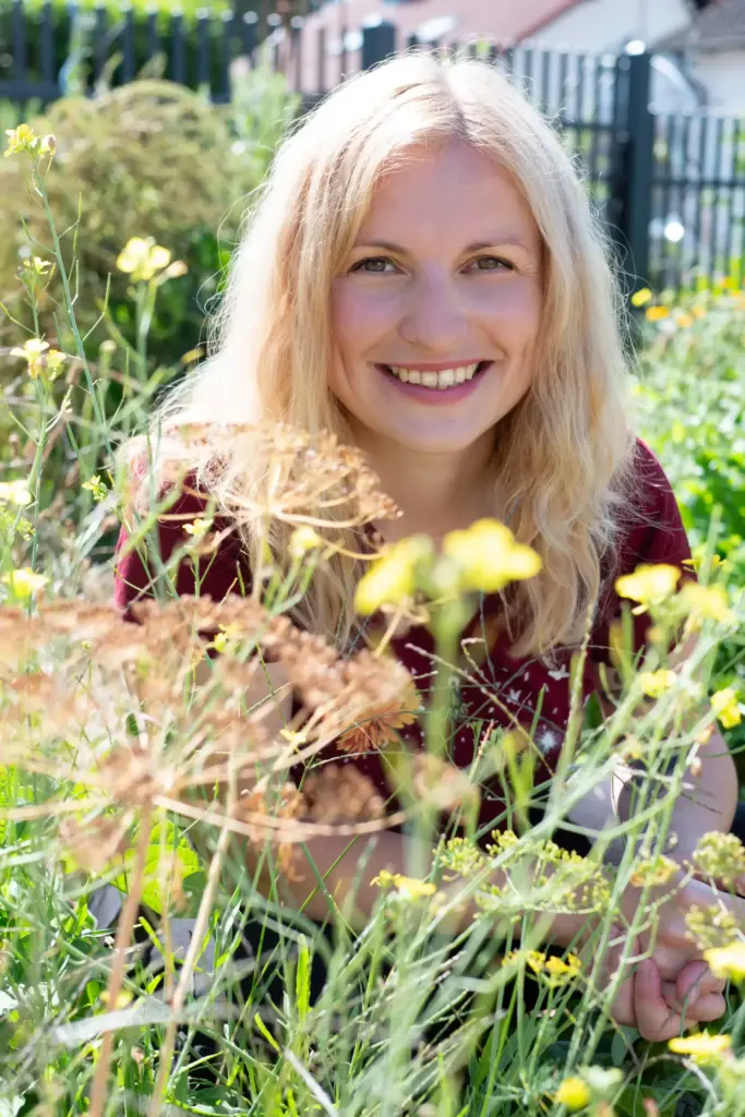 Lächelnde Frau in einem blühenden Garten, umgeben von Kräutern und Wildblumen, bei der nachhaltigen Beratung für Garten und Balkon. Neben der Gartenberatung bietet Julia zudem Veranstaltungen in Form von Kräuterwanderungen im Saarland, Kindergeburtstag, Junggesellenabschied und Workshops für Selbstversorger an, die in der Natur stattfinden.